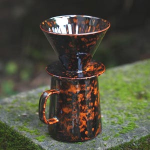 Peut inclure: Un ensemble de cafetière à verser en céramique noire et orange mouchetée. L'ensemble comprend un verseur conique, un couvercle et une tasse avec une anse. La cafetière est posée sur une surface en pierre moussue.