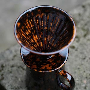 K&ouml;nnte beinhalten: Ein Keramik-Kaffee-Pour-Over-Set mit dunkelbrauner und orangefarbener gesprenkelter Glasur. Das Set enth&auml;lt einen Dripper und eine Tasse mit Henkel, perfekt f&uuml;r die Kaffeezubereitung zu Hause. Das Design ist sowohl funktional als auch stilvoll.