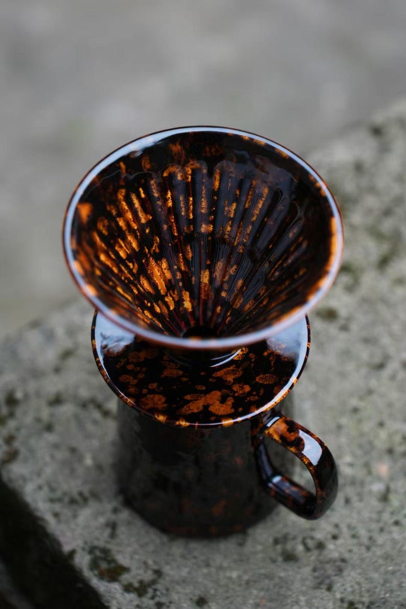 K&ouml;nnte beinhalten: Ein Keramik-Kaffee-Pour-Over-Set mit dunkelbrauner und orangefarbener gesprenkelter Glasur. Das Set enth&auml;lt einen Dripper und eine Tasse mit Henkel, perfekt f&uuml;r die Kaffeezubereitung zu Hause. Das Design ist sowohl funktional als auch stilvoll.