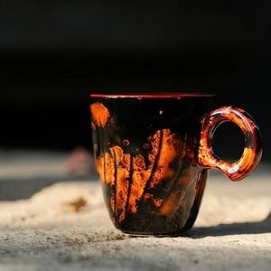 May include: A ceramic mug with a black exterior and orange, lava-like patterns. The rim is red, and the handle is a matching orange. The mug is sitting on a light-colored surface.
