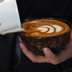 May include: A hand-held, brown ceramic bowl filled with coffee, featuring a white and brown latte art design. A white pitcher pours milk into the coffee, creating a swirling pattern. The bowl has a dark, speckled exterior.