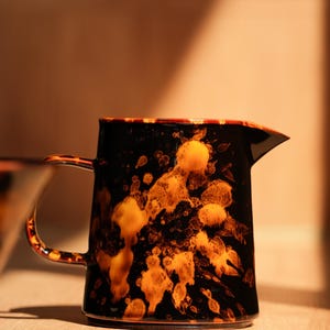 K&ouml;nnte beinhalten: Eine schwarz-orangefarbene Keramikkrug mit Henkel und Ausguss. Der Krug hat ein gesprenkeltes Design mit orangefarbenen und gelben Akzenten. Der Griff und der Rand sind ebenfalls orange. Der Krug steht auf einer Holzoberfl&auml;che.
