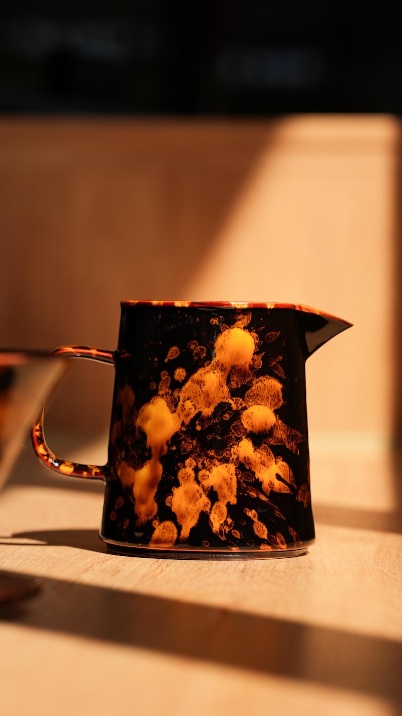 K&ouml;nnte beinhalten: Eine schwarz-orangefarbene Keramikkrug mit Henkel und Ausguss. Der Krug hat ein gesprenkeltes Design mit orangefarbenen und gelben Akzenten. Der Griff und der Rand sind ebenfalls orange. Der Krug steht auf einer Holzoberfl&auml;che.