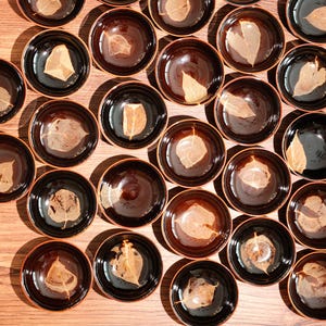 May include: Overhead shot of numerous small, round, brown and black bowls, each containing a dried leaf. The bowls are arranged on a light brown wooden surface, creating a visually appealing pattern. The leaves vary in shape and color.