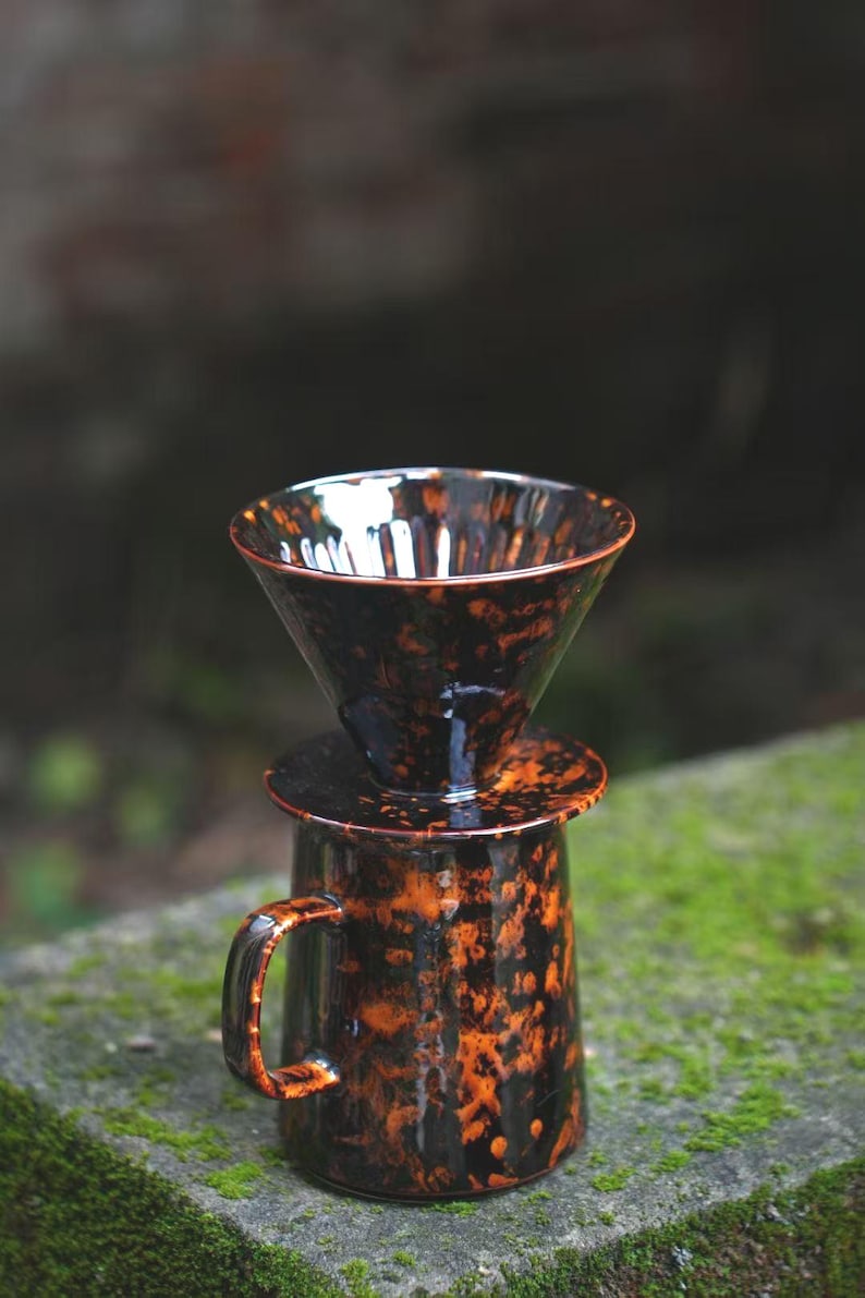 K&ouml;nnte beinhalten: Ein Kaffeebereiter-Set aus Keramik mit gesprenkelter schwarz-oranger Glasur. Das Set besteht aus einem konischen Dripper, einem Deckel und einer Tasse mit Henkel. Der Kaffeebereiter steht auf einer moosigen Steinoberfl&auml;che.