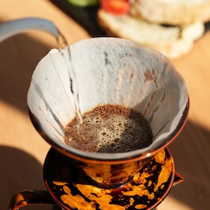 K&ouml;nnte beinhalten: Nahaufnahme eines Kaffee-Pour-Over-Setups. Ein braun-oranger Keramik-Pour-Over-Kaffeebereiter mit Papierfilter wird mit hei&szlig;em Wasser gef&uuml;llt. Gurkenscheiben, Tomaten und Brot im Hintergrund.