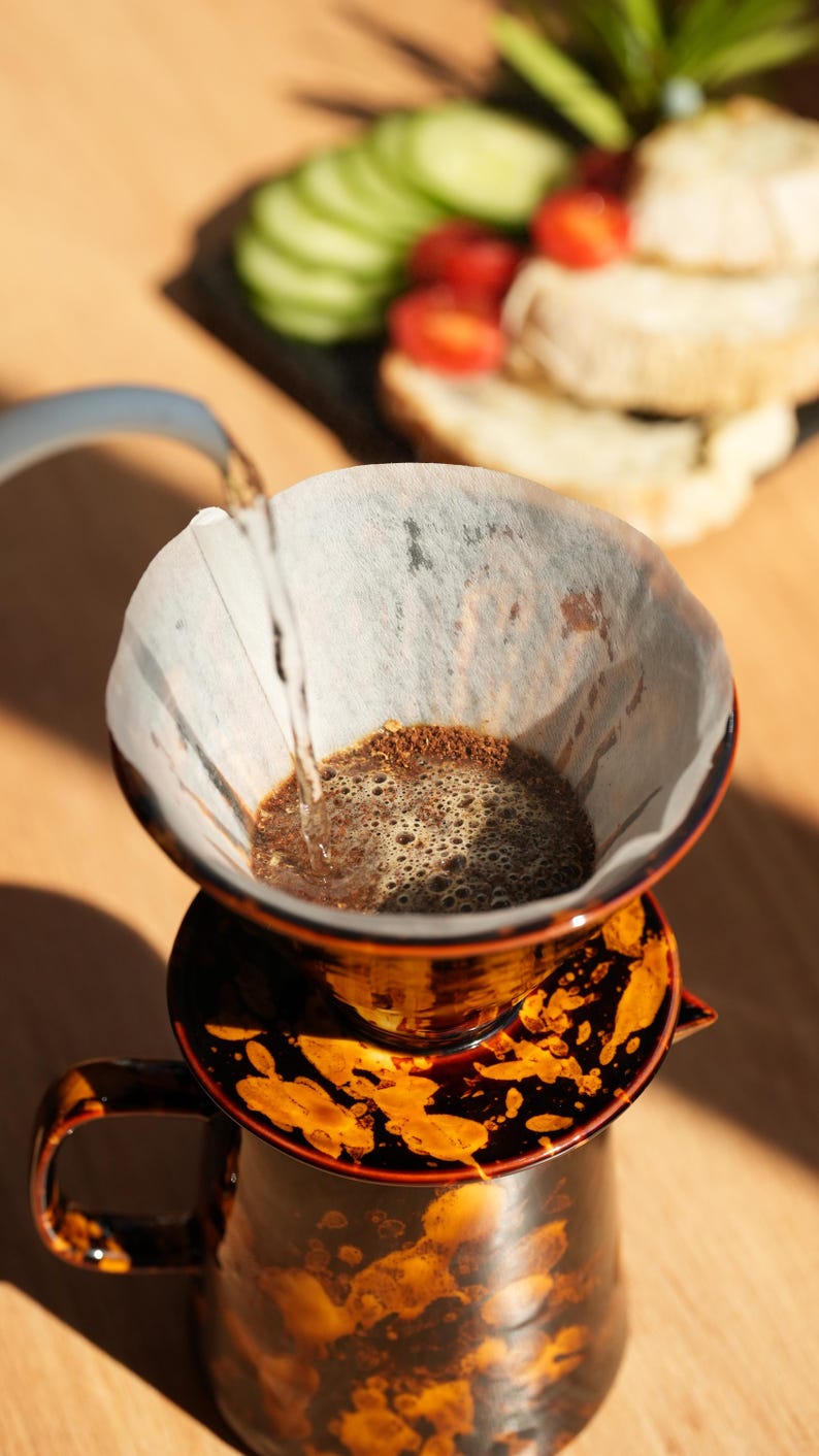K&ouml;nnte beinhalten: Nahaufnahme eines Kaffee-Pour-Over-Setups. Ein braun-oranger Keramik-Pour-Over-Kaffeebereiter mit Papierfilter wird mit hei&szlig;em Wasser gef&uuml;llt. Gurkenscheiben, Tomaten und Brot im Hintergrund.