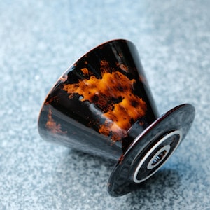 K&ouml;nnte beinhalten: Ein schwarzer Keramik-Kaffeefilter mit orangefarbenen und braunen Mustern. Der Filter hat eine konische Form und einen kreisf&ouml;rmigen Sockel mit einem silbernen Ring. Das Design ist abgewinkelt und zeigt das Innere des Filters.
