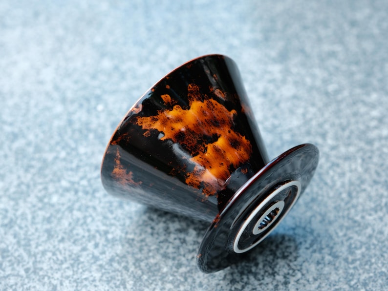K&ouml;nnte beinhalten: Ein schwarzer Keramik-Kaffeefilter mit orangefarbenen und braunen Mustern. Der Filter hat eine konische Form und einen kreisf&ouml;rmigen Sockel mit einem silbernen Ring. Das Design ist abgewinkelt und zeigt das Innere des Filters.