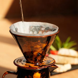 K&ouml;nnte beinhalten: Ein Keramik-Pour-Over-Kaffeebereiter mit braun-orangefarbener, gesprenkelter Glasur. Hei&szlig;es Wasser wird in den Filterkonus gegossen. Die Kaffeekanne steht auf einer Holzoberfl&auml;che, was auf einen Hausbrauaufbau hindeutet.