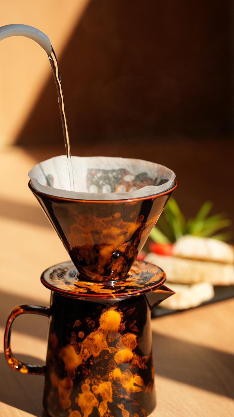 K&ouml;nnte beinhalten: Ein Keramik-Pour-Over-Kaffeebereiter mit braun-orangefarbener, gesprenkelter Glasur. Hei&szlig;es Wasser wird in den Filterkonus gegossen. Die Kaffeekanne steht auf einer Holzoberfl&auml;che, was auf einen Hausbrauaufbau hindeutet.