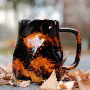 May include: A black ceramic mug with an organic shape and a large handle. The mug has a glossy finish with orange and brown splatter-like patterns. The mug is surrounded by fallen leaves.