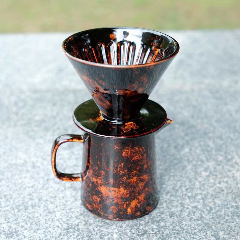 K&ouml;nnte beinhalten: Ein Keramik-Pour-Over-Kaffeezubereiter-Set. Das Set beinhaltet eine dunkelbraune, gesprenkelte Kaffeekanne mit Griff, einen passenden Deckel und einen konisch geformten Kaffeetropfer. Der Tropfer hat vertikale Rillen im Inneren.