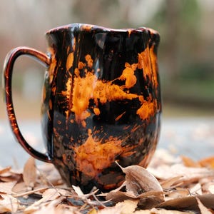 May include: A handmade ceramic mug with a unique design. The mug features a black base with orange splatter patterns, a curved handle, and a slightly wavy rim. The mug is sitting on a surface covered with fallen leaves.