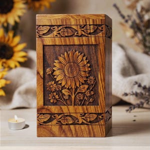 Op de afbeelding: Een houten urn met een gebeeldhouwd zonnebloemontwerp. De rechthoekige doos is gemaakt van donkerbruin hout, met een gedetailleerd zonnebloem- en bloemenpatroon dat in het voorpaneel is gesneden. De boven- en onderranden hebben een gebeeldhouwd bladontwerp.