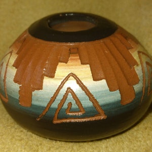 May include: A round, ceramic vessel with a dark brown base and a black rim. The body features a geometric design in shades of brown, tan, and blue. A triangle with a spiral is etched into the front.