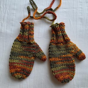 Op de afbeelding: Gebreide wanten in oranje, groen en bruin, verbonden door een bijpassend koord. De wanten hebben een geribbelde manchet en een getextureerd uiterlijk, perfect voor de winter.