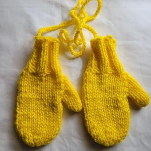Puede incluir: Un par de mitones de punto de color amarillo brillante con un largo cordel amarillo para atar. Los mitones están hechos con un punto de tejido sencillo.