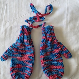 Pode incluir: Um par de luvas de malha em azul e vermelho. As luvas são conectadas por um cordão de malha. O padrão de cores são listras horizontais azuis e vermelhas. As luvas estão sobre uma superfície branca.