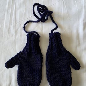 May include: A pair of navy blue knitted mittens connected by a matching cord. The mittens are made of a thick yarn, with a ribbed cuff and a rounded shape. The mittens are lying on a white surface.