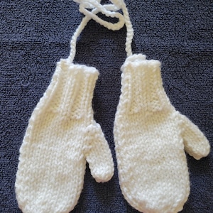 Puede incluir: Un par de guantes de punto blanco con un lazo de lana blanco. Los guantes están hechos con un punto de tejido simple y tienen una forma clásica de guante.