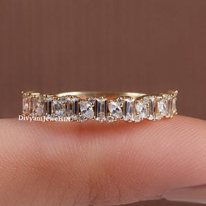 May include: A gold band ring with a continuous row of alternating rectangular and oval-shaped clear gemstones. The ring is displayed on a finger against a blurred brown background. The text "DivyamjewelsIN" is visible on the ring.