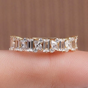 May include: A gold band ring with a continuous pattern of clear, rectangular and square-cut gemstones. The ring is set with multiple stones, creating a sparkling effect. The text "DivyamJewels" is visible on the ring.