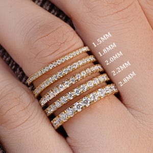 May include: Six gold-colored eternity rings with clear gemstones are displayed on a finger. The rings vary in size, with the text indicating widths of 1.5mm, 1.8mm, 2.0mm, 2.2mm, and 2.8mm. The rings are set with small, round gemstones.