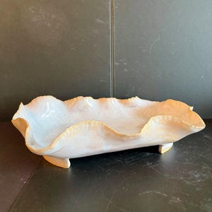May include: An elongated, rectangular ceramic tray with a wavy, ruffled rim. The piece features a glossy, off-white glaze with subtle peach and tan gradients along the edges. It is supported by four small, integrated feet at the base.