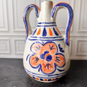 May include: A white ceramic vase with two handles, adorned with a vibrant floral design in orange and blue. The vase features blue handles with orange accents and decorative blue and orange patterns.