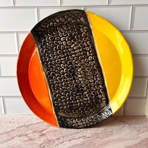 May include: A decorative ceramic plate with a unique design. The plate features a black, textured center section with a pattern of small, oval shapes. The plate is divided into sections of orange and yellow.