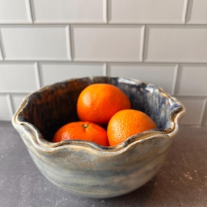 May include: A ceramic bowl with a wavy rim, glazed in shades of blue and brown, holds three bright orange tangerines. The bowl sits on a dark gray surface, with a white tile backsplash in the background. The bowl is a decorative kitchen item.