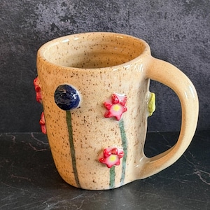 May include: A ceramic mug with a speckled beige glaze featuring raised, three-dimensional floral decorations. The mug displays small painted flowers in red, yellow, and blue with green stems extending down the side of the cylindrical body.