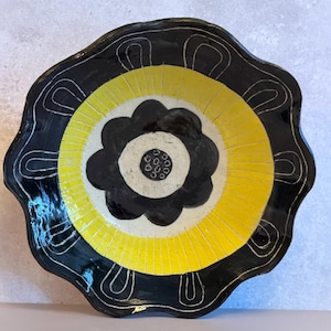 May include: A round, scalloped-edge ceramic bowl featuring a hand-painted design. The center displays a black flower on a white background, surrounded by a bright yellow ring with radiating lines, and a black border with white line motifs.