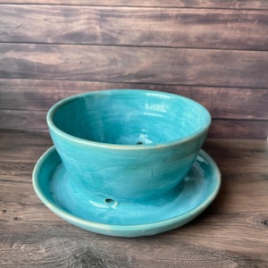 May include: A turquoise ceramic planter with a matching saucer. The planter has a rounded bowl shape with drainage holes, and the saucer is a shallow dish. The set is glazed with a glossy finish, and the color is a vibrant teal.