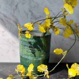 May include: A green ceramic vase displays yellow flowers with brown markings. The vase has a textured finish and a cylindrical form. The flowers are arranged to create a graceful display, offering a striking contrast.