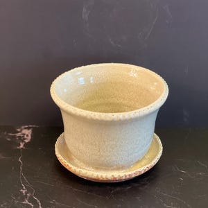 May include: A cream-colored ceramic planter with a matching saucer. The planter has a textured surface and a decorative rim. The saucer has a contrasting orange and brown trim. The planter and saucer are on a black surface.