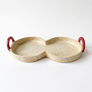 May include: A double-sectioned ceramic tray with a speckled cream and beige finish. The tray features two rounded compartments joined in the center and two bright red, curved handles attached to the outer edges of each side.