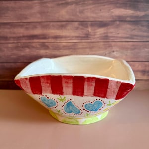 May include: A square ceramic bowl with a white interior and a hand-painted exterior. The rim features vertical red stripes, while the body displays a pattern of light blue hearts, small green star-like accents, and a light green base.