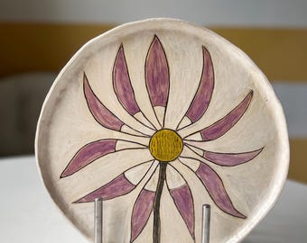 May include: A ceramic plate with a hand-painted floral design. The plate features a cream-colored background with a large purple and yellow flower in the center. The flower has purple petals and a yellow center. The plate is displayed on a metal stand.