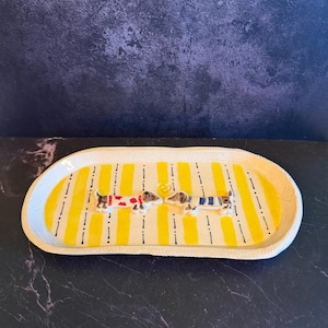 May include: An oval ceramic tray with a speckled cream base featuring vertical yellow stripes and thin black dashed lines. Two sculpted dachshund figures are attached to the center, one wearing a red patterned sweater and one in a blue striped sweater.