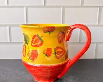 May include: A ceramic mug with a bright yellow upper half and a red base and handle. The yellow section is decorated with red strawberries and green leaves. The mug is designed for beverages.