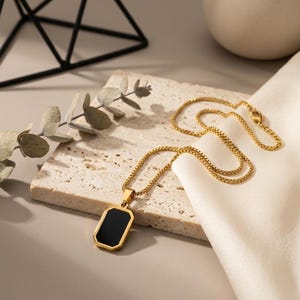 Op de afbeelding: Een goudkleurige box chain ketting met een rechthoekige hanger voorzien van een gladde, effen zwarte inleg. De hanger heeft een gepolijst goudkleurig metalen frame met afgeschuinde randen en de ketting bestaat uit kleine, uniforme vierkante schakels.