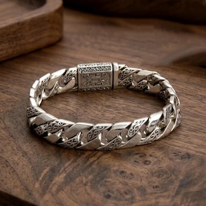 Puede incluir: Una pulsera de plata con un diseño de cadena de eslabones. La pulsera presenta un cierre adornado y detalles decorativos en cada eslabón. La joya se exhibe sobre una superficie de madera.