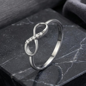 Peut inclure: Une bague argentée présentant un motif de symbole infini sur l'anneau. La boucle de l'infini est ornée de quatre petites pierres rondes et transparentes serties sur un côté. L'intérieur de l'anneau porte l'inscription S925.