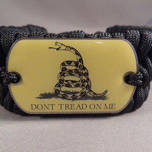 May include: A black paracord bracelet with a metal tag featuring a yellow background and a black and white image of a coiled snake with the text "Don't Tread On Me".