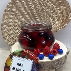 Könnte beinhalten: Ein Kerzenglas mit Wildbeeren-Marmeladen-Motiv. Die Kerze ist rot und schwarz, in einem durchsichtigen Glas. Der Deckel trägt den Text "WILD BERRY JAM". Dekorative Wachsstücke in Beerenform sind um die Kerze herum angeordnet.