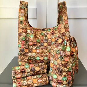 May include: A set of three reusable shopping bags made of fabric with a vintage bottle cap print. The bags are brown, orange, and yellow with a variety of bottle cap designs. The bags are folded and displayed on a gray surface.