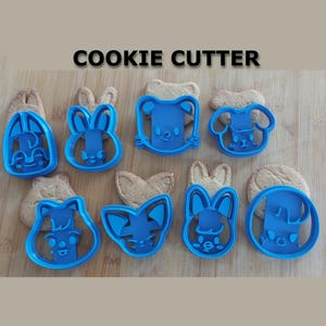Peut inclure: Plusieurs découpoirs à biscuits en plastique bleu en forme d'animaux, dont des lapins, des chiens et des ours, sont présentés sur une surface en bois. Le mot "COOKIE CUTTER" est imprimé en noir au-dessus. Les découpoirs sont placés sur des biscuits.
