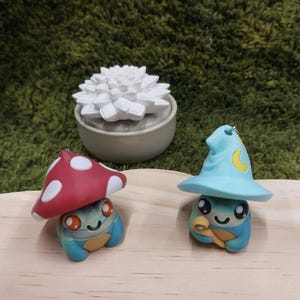 Op de afbeelding: Twee grillige kleifiguren: een met een rode paddenstoelkap en de andere met een blauwe tovenaars hoed. Een witte vetplant staat in een grijze keramische pot. De beeldjes zijn blauw en bruin met lachende gezichten. De achtergrond is groen.